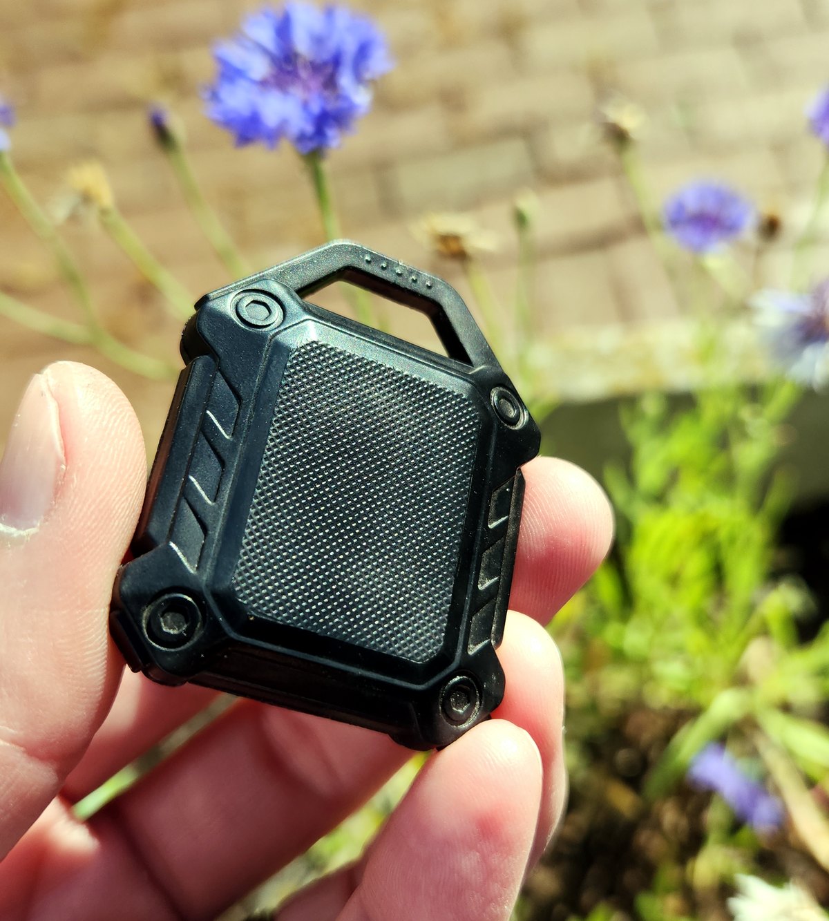 A rugged black clip-on case for an AirTag, with a carabiner loop, held in a hand outdoors with cornflowers behind