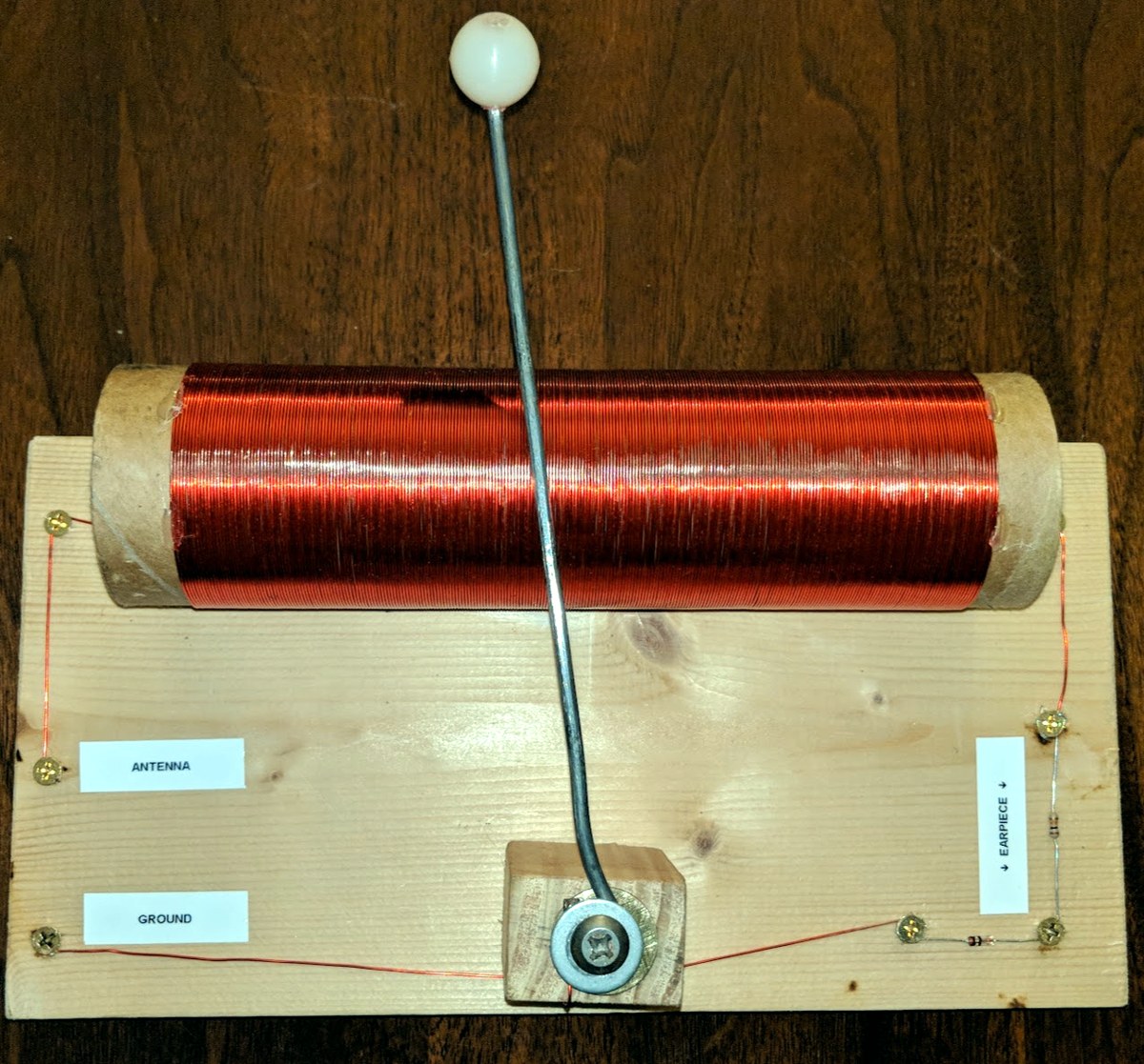A homemade crystal radio: a large copper coil on a cardboard tube mounted on a wooden base, with a metal slider arm for tuning, labeled antenna and ground terminals, and a diode wired to earpiece terminals