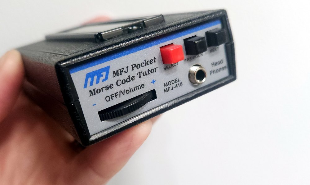 An MFJ Pocket Morse Code Tutor, model MFJ-418, held in a hand - a small handheld device with a volume dial, a red select button, previous/next buttons, and a headphone jack