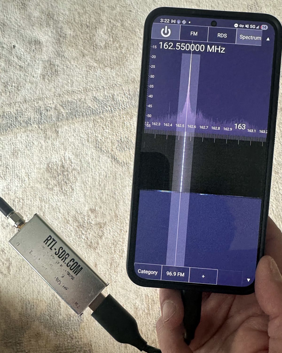 An RTL-SDR dongle plugged into a phone, its screen showing the 162.550 MHz NOAA weather channel with a strong signal spike