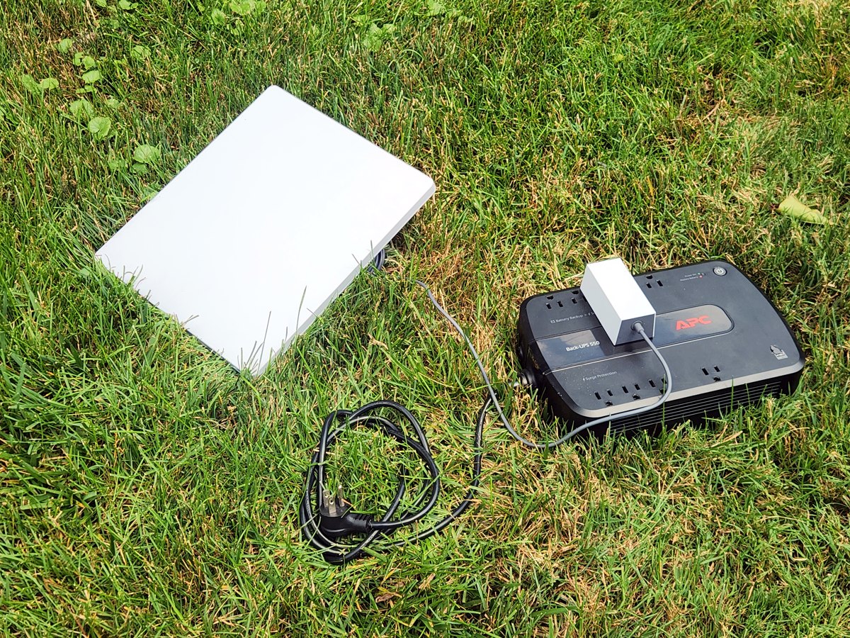 A Starlink Mini dish lying on grass, its power cable running to a black APC Back-UPS 550 unit, whose own wall plug is coiled and unplugged on the ground - the dish running entirely off the UPS battery, off-grid