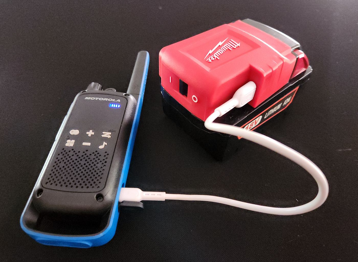 An 18V tool battery with a USB adapter charging a handheld two-way radio